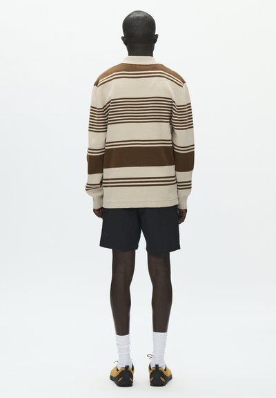 Pull à manches longues à rayures beige et marron, porté sur un short noir. Le pull présente des rayures horizontales de largeurs variées.