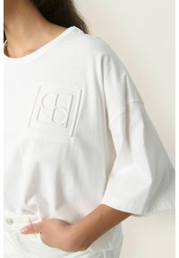 Hvid bomulds oversized t-shirt med et tekstureret logo på brystet, korte ærmer og en bred udskæring. Glat stof med afslappet pasform.
