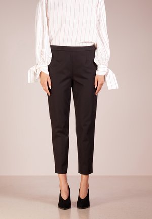 Kvinde iført sorte, tilspidsede ankellange bukser, hvid bluse med røde striber og bundne manchetter samt sorte højhælede sko, stående mod en ensfarvet baggrund.