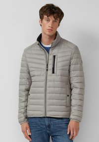 Lichtgrijze gewatteerde jas met ritssluiting, hoge kraag, borstzak aan de voorkant en zijzakken. Zachte textuur met horizontale doorgestikte lijnen.