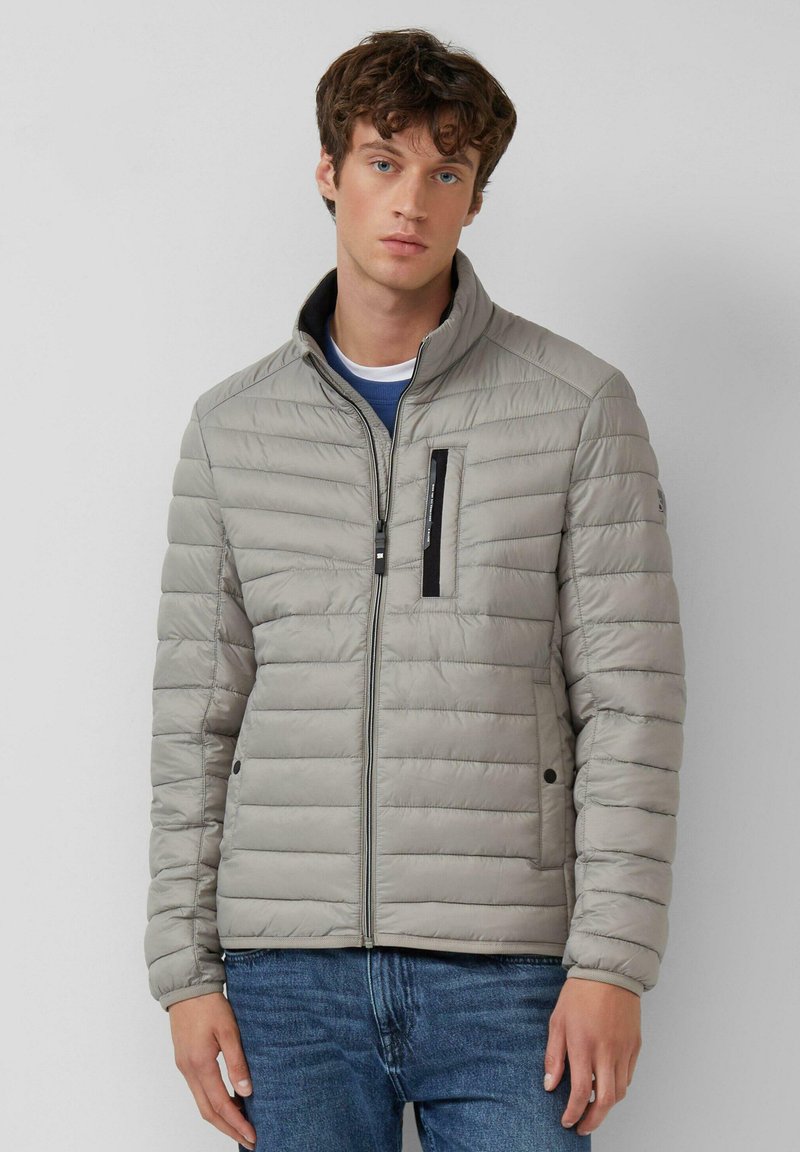 Lichtgrijze gewatteerde jas met ritssluiting, hoge kraag, borstzak aan de voorkant en zijzakken. Zachte textuur met horizontale doorgestikte lijnen.
