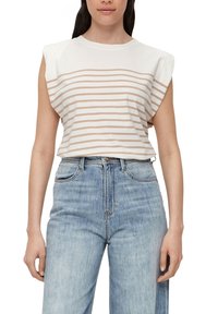 Žena v bezrukávovém bílém topu s béžovými vodorovnými pruhy a světle modrých džínech se zvýšeným pasem, stojící na bílém pozadí.