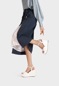 Sandali wedge bianchi con suola piatta, punta aperta e cinturino alla caviglia. L'outfit include una blusa azzurro chiaro e una gonna blu scuro.