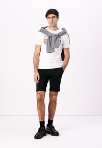 T-shirt grafica bianca, maglione grigio appoggiato sulle spalle, pantaloni corti neri, scarpe nere, texture a contrasto, design minimalista.