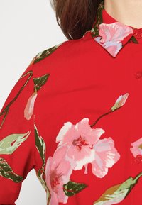 Camicia rossa con motivo floreale con accenti rosa e verdi, colletto e dettagli con bottoni sul davanti.