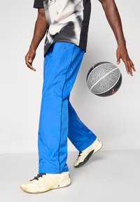 Blauwe sportbroek met een gladde textuur en zijpiping, gecombineerd met een gestreepte basketbal en beige sportschoenen.