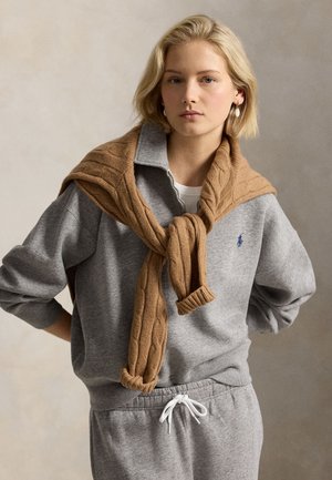 Jeune femme aux cheveux blonds courts, portant un sweat gris et un pantalon de survêtement, un pull marron posé sur les épaules, debout les mains sur les hanches.