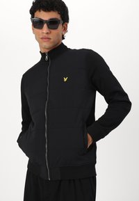 Giacca nera con zip, frontale imbottito, maniche lunghe, polsini e orlo a costine. Presenta un piccolo logo giallo sul petto.