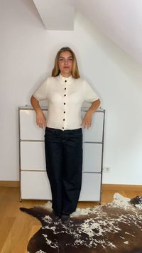 Weiße kurzärmlige Strickjacke mit schwarzen Knöpfen und einem gerippten Muster, kombiniert mit dunkelblauen weit geschnittenen Jeans, vor einem neutralen Hintergrund.