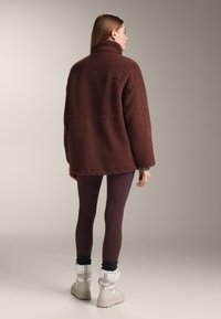 Hnědá fleecová bunda s vysokým límcem, texturovaná látka a volný střih, doplněná fialovými legínami a krémovými sněhovými botami.