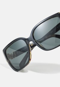 Ochelari de soare supradimensionați negri cu lentile gri închis, având un accent de scoică pe ramă și detalii gravate ale mărcii pe tâmplă.
