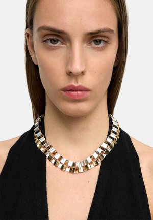 Kobieta ubrana w czarny top na szyję oraz masywny metaliczny naszyjnik z naprzemiennie ułożonymi złotymi i srebrnymi prostokątnymi ogniwami.