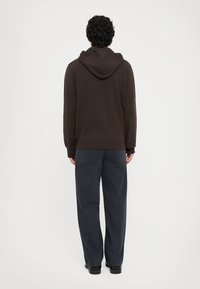 Brown hoodie with a drawstring hood, ribbed cuffs and hem. Paired with dark blue, wide-leg trousers. Model facing away from the camera.