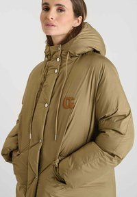 Olijfgroene gewatteerde jas met een hoge kraag, metalen drukknoopsluiting, elastische trekkoorden en een bruine logo patch op de borst.