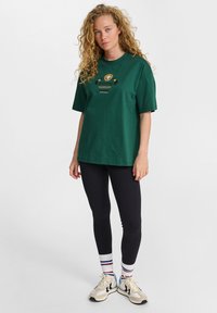 Zielona bawełniana koszulka z luźnym krojem, z graficznym wzorem na piersi, w połączeniu z czarnymi legginsami i skarpetkami w wielokolorowe paski.