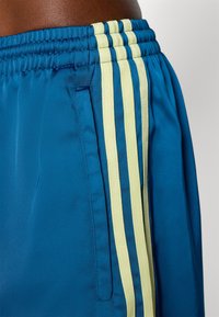 Shorts de sport bleus avec une texture lisse, arborant trois bandes latérales jaunes. Taille élastique avec un détail de poche unique.