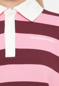 Pruhovaná polo košieľka v marhuľových a ružových farbách, s bielym golierom a logom "COLOURFUL REBEL" vyšitým bielou niťou.