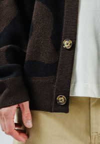 Braune Strickjacke mit dunklem Camouflage-Muster, gerippten Bündchen und Knopfdetail mit Vierloch-Design. Wird über einem hellen Oberteil getragen.