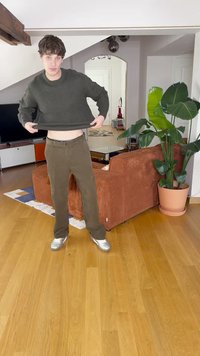 Zaļš adīts džemperis, īsa dizaina, apvienots ar brūnām biksēm. Sudraba sneakers, koka grīda un pods ar augu redzami fona attēlā.