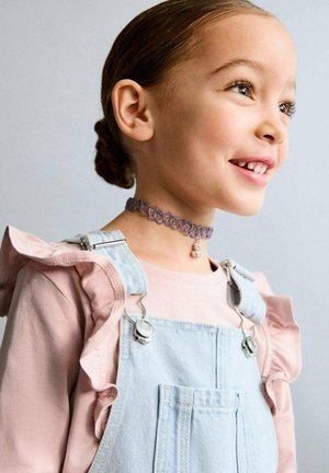 Chemise rose clair avec manches à volants et salopette en denim avec ferrures argentées ; un collier ras du cou violet avec un pendentif ours autour du cou.