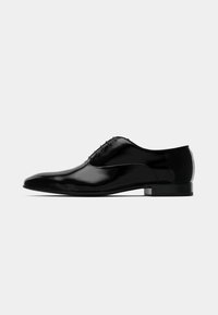 Elegante scarpa da uomo in pelle nera lucida, con allacciatura chiusa e tacco basso, mostrata in profilo laterale su uno sfondo bianco semplice.