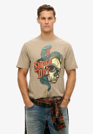 T-shirt en coton beige avec un graphique coloré de crâne et de serpent, portant le texte "SUPER DRY". Associé à un jean en denim bleu et une chemise à carreaux nouée à la taille.
