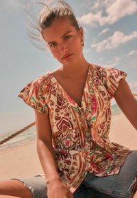 Chemise à motifs floraux avec un col en V, des manches courtes à volants et des teintes multicolores de rose, vert et beige sur un tissu texturé.