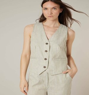Femme aux longs cheveux bruns portant un gilet sans manches beige boutonné et un pantalon assorti à rayures blanches verticales, sur un fond uni.