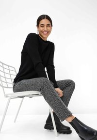 Schwarzer gerippter Pullover, graue Fischgrätenhosen und schwarze klobige Stiefeletten. Model sitzt auf einem weißen Metallstuhl vor einem schlichten Hintergrund.