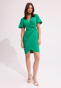 Zielona sukienka o kroju kopertowym z krótkimi falbaniastymi rękawami, skręconym detalem w pasie i asymetrycznym dołem do kolan. Gładki materiał.