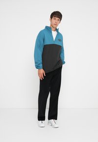 Ung mann står mot hvit bakgrunn iført blå og svart quarter-zip genser, svarte bukser og hvite sneakers.