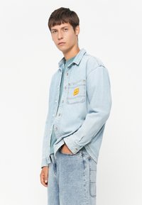 Light blue denim shirt with a button-up front, one chest pocket featuring a yellow label, and rolled sleeves. Worn over a teal top.
