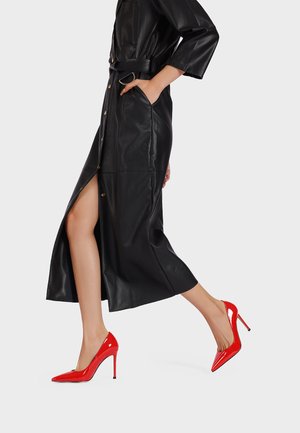 Tacco alto in pelle verniciata rossa, con punta affilata e tacco a stiletto, indossati con un lungo abito in pelle nera caratterizzato da una apertura laterale e tasche.