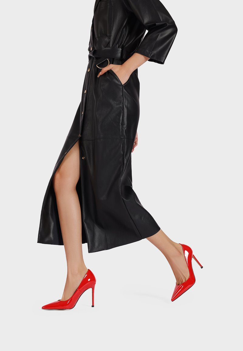 Tacco alto in pelle verniciata rossa, con punta affilata e tacco a stiletto, indossati con un lungo abito in pelle nera caratterizzato da una apertura laterale e tasche.