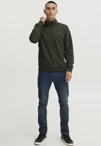 Sudadera de media cremallera de color verde oscuro con cuello, que presenta un pequeño logo en el pecho, combinada con vaqueros azules y zapatillas negras.