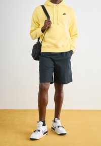 Felpa gialla con logo nero, pantaloni corti neri con tasche laterali e sneakers bianche con accenti neri. Ambientato su un pavimento giallo.