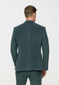 Tölgyzöld kordbársony blézer vertikális bordás textúrával. Jellemzője a testreszabott fazon, hátsó részen egyetlen szellőzőnyílás és gombos mandzsetta részletek.