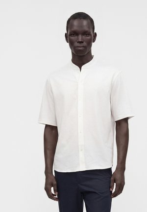 Homme adulte portant une chemise blanche à manches courtes avec un col mandarin et un pantalon foncé, debout devant un fond blanc uni.