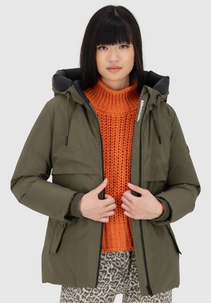 Veste isolée vert olive avec une capuche noire, fermeture éclair, et deux poches avant, superposée sur un pull en laine orange et un pantalon à motifs.