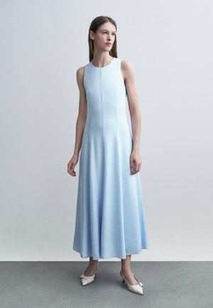 Γυναίκα που στέκεται φορώντας αμάνικο, ανοιχτό μπλε φόρεμα μέχρι τον αστράγαλο και λευκές μυτερές γόβες, μπροστά σε απλό γκρι φόντο.