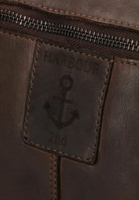 Etichetta in pelle marrone con un design ad ancoraggio impresso, con il testo "HARBOUR" e "2nd". I bordi cuciti conferiscono texture.