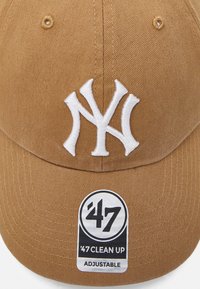 Braune Baumwollmütze mit einem gestickten weißen "NY"-Logo, einem gebogenen Schirm und einem verstellbaren Riemen mit einem schwarz-weißen Etikett.