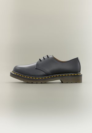 Schwarzer glatter Lederschnürschuh mit gelber Naht und dicker Gummisohle, seitlich auf neutralem Hintergrund abgebildet.