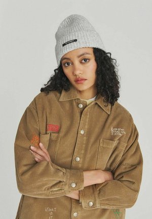 Giovane donna con capelli ricci indossa un berretto grigio di maglia e una giacca oversize in velluto a coste color cammello con testo ricamato, braccia incrociate, sfondo neutro.