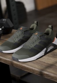 Paire de chaussures de sport vertes et noires avec semelles blanches sur un banc en bois, poids de gym flous en arrière-plan.