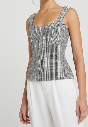 Kobieta ubrana w dopasowany, czarno-biały top w kratę bez rękawów z szerokimi ramiączkami, zestawiony z luźnymi białymi spodniami.