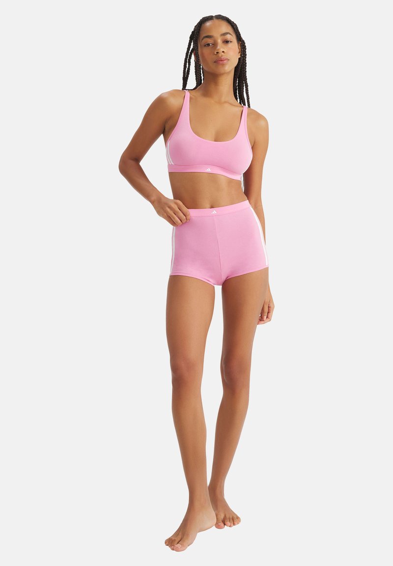 Frau steht barfuß und trägt einen pinkfarbenen Sport-BH und passende hoch taillierte Shorts mit weißen Seitenstreifen vor einem schlichten weißen Hintergrund.