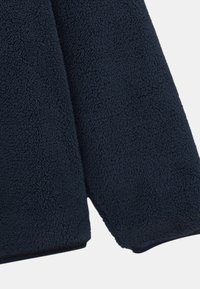 Bords de manches d'une veste en polaire bleu foncé avec une surface douce, texturée et duveteuse sur un fond clair.