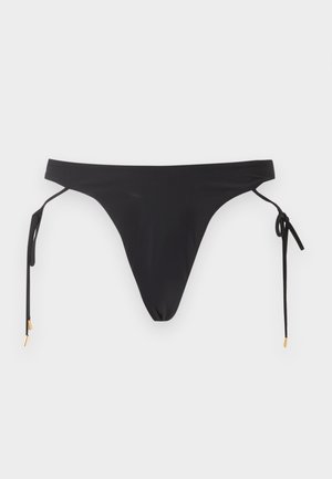 Kobieta stojąca boso, ubrana w top bikini w kolorze rdzy i czarne majtki bikini z wiązaniami po bokach na białym tle.