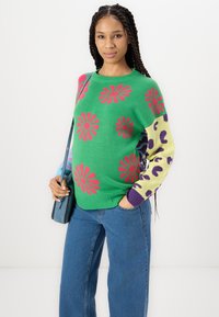 Πράσινο πλεκτό πουλόβερ με ροζ λουλουδάτα μοτίβα, αντίθετους πολύχρωμους μανίκους με λεοπάρ εκτύπωση, συνδυασμένο με μπλε τζιν και τσάντα ώμου.